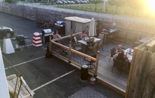 Outdoor dining area with wooden deck, tables, chairs, umbrellas, and guests eating.