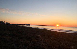 Serene beach sunrise with the sun near the horizon, calm waters, and a distant pier.