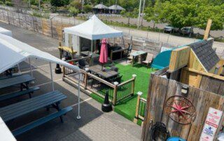 Outdoor seating area with picnic tables, tents, and fenced spaces on a sunny day.