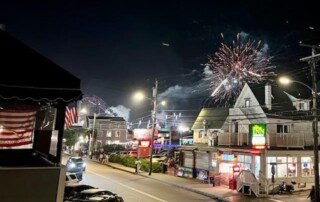 Street view at night with fireworks in the sky, buildings, and parked cars visible.